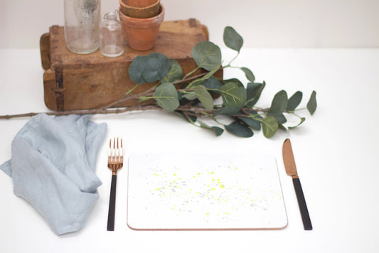 A yellow splatter placemat with a small blue napkin and a brown fork, set on a white surface with a few green leaves and a pot in the background.