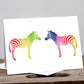 A greeting card featuring two stylized, rainbow-colored zebras connected nose to nose on a white background, displayed on a wooden surface.