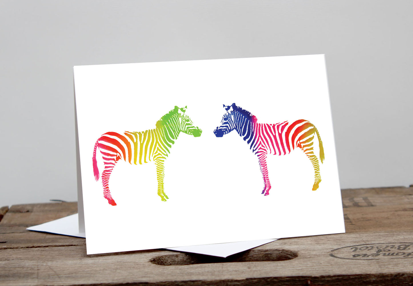 A greeting card featuring two stylized, rainbow-colored zebras connected nose to nose on a white background, displayed on a wooden surface.