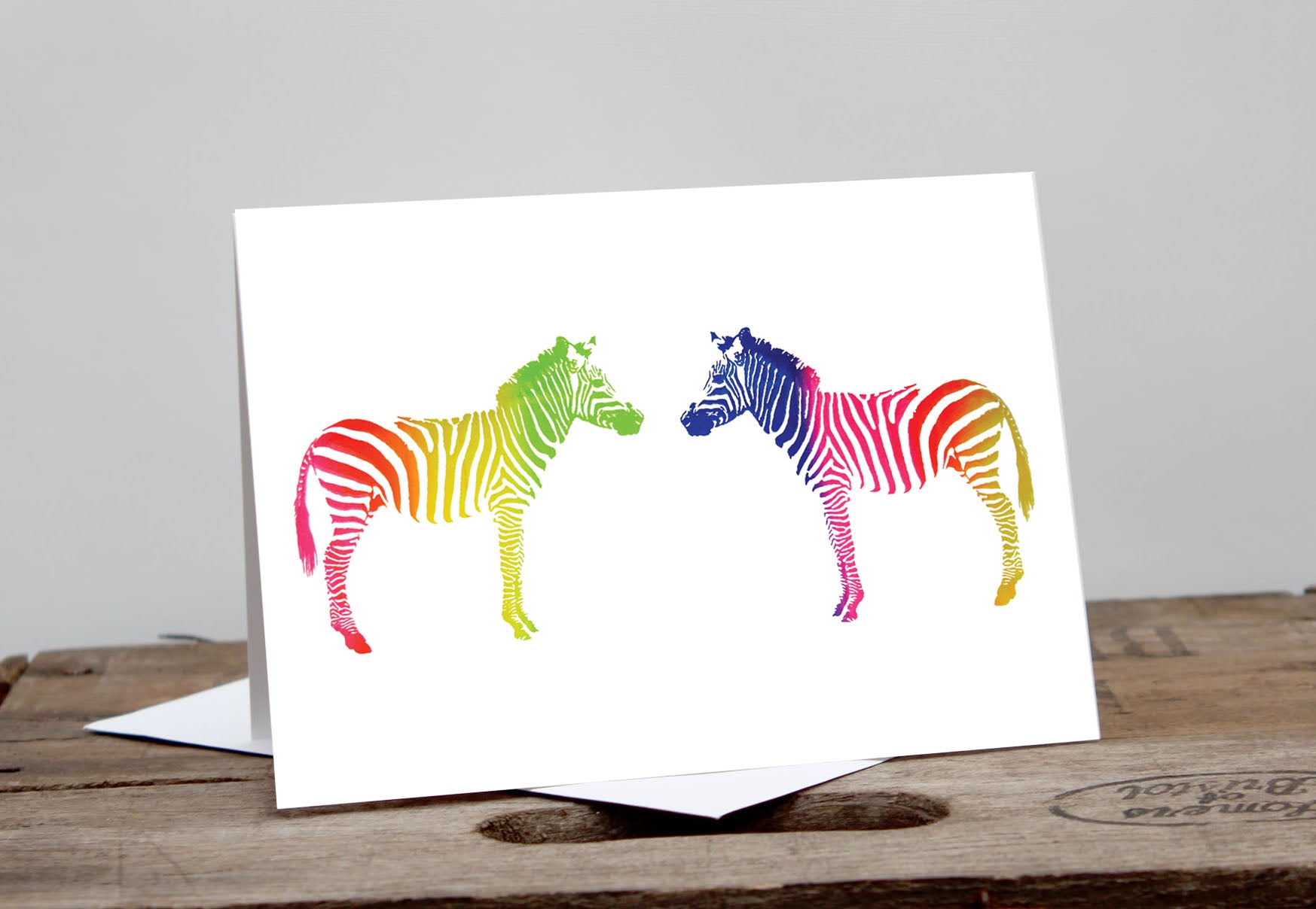 A greeting card featuring two stylized, rainbow-colored zebras connected nose to nose on a white background, displayed on a wooden surface.